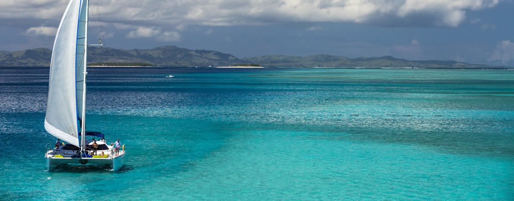 Velero a lo lejos sobre aguas de color turquesa y azul en Fiji